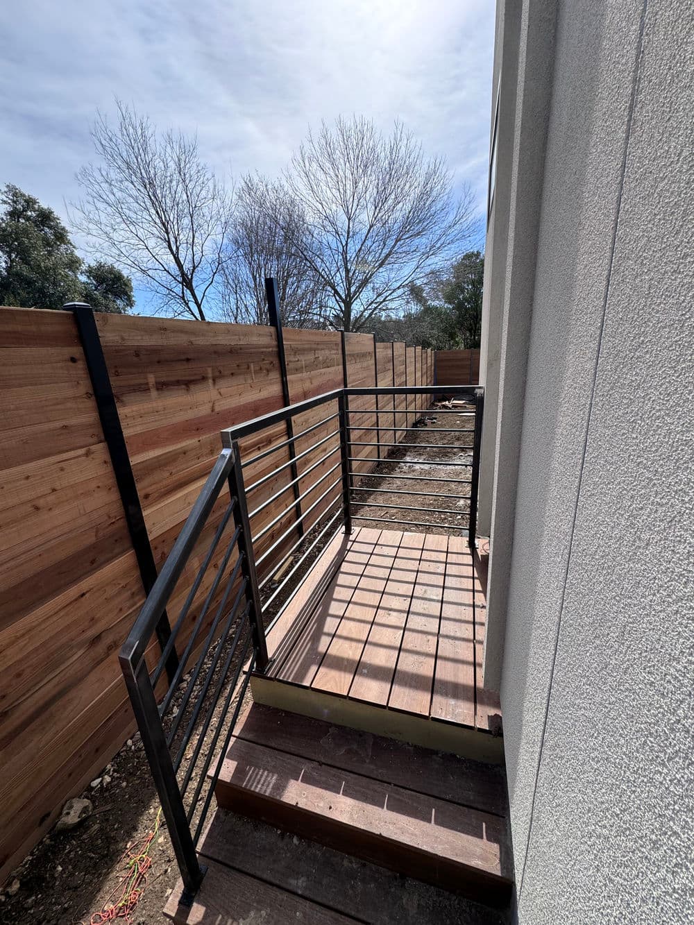 Modern outdoor staircase with metal railing beside wooden fence and bare trees. Building exterior.