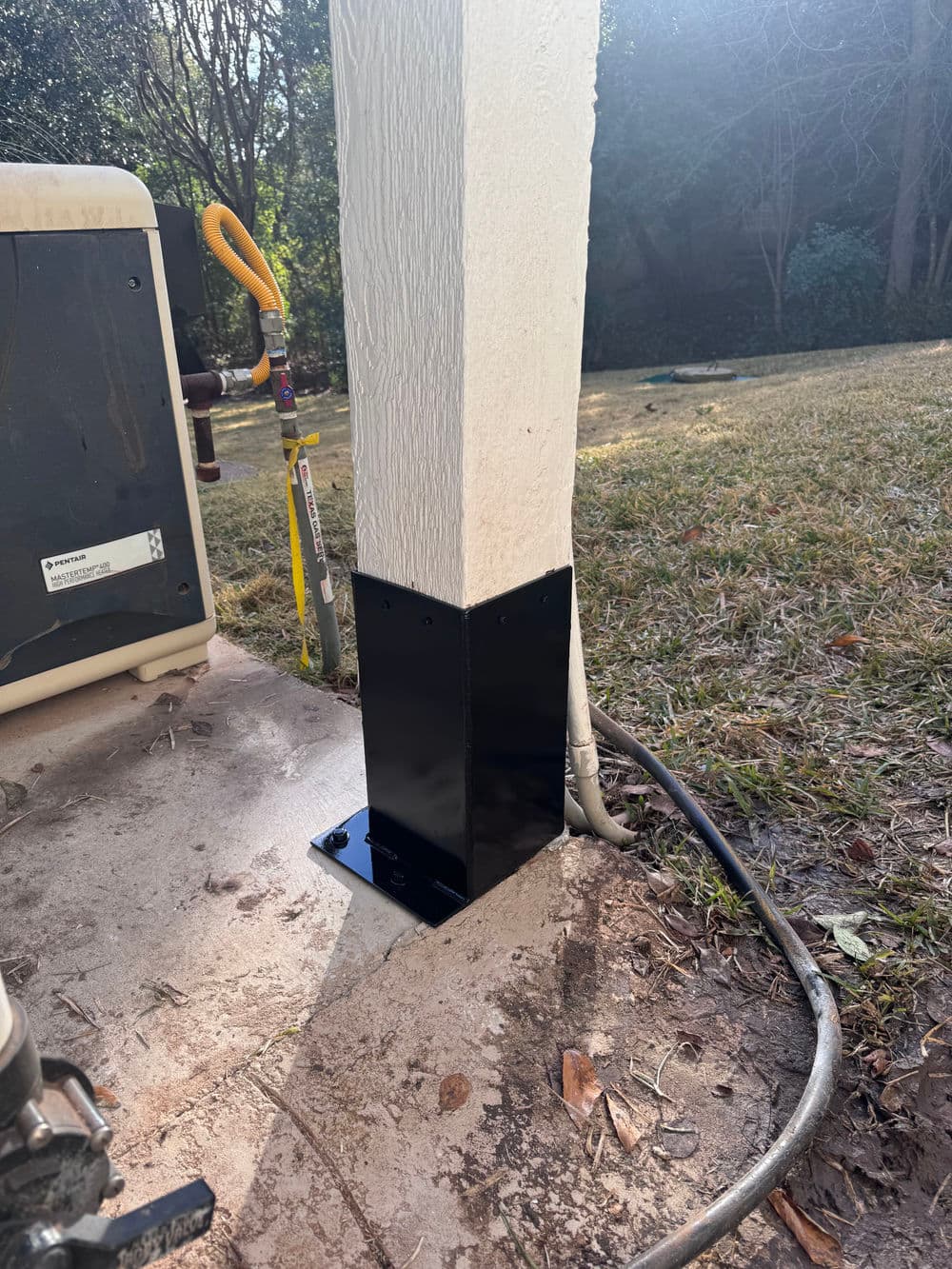 Black metal post base securing a white outdoor column near HVAC unit and grassy area.