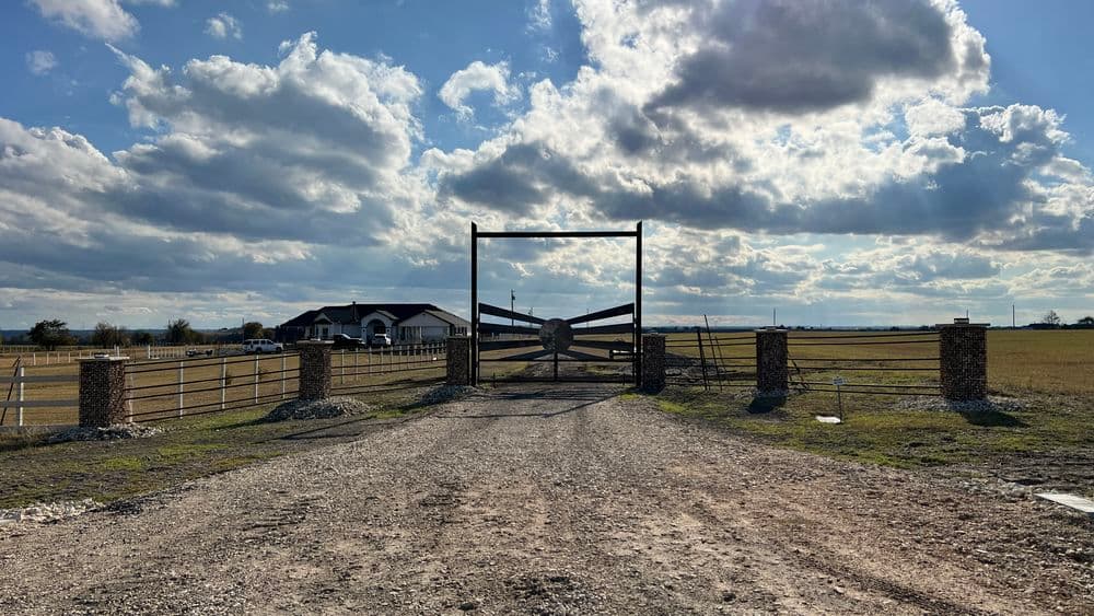 Rural entrance gate with cloudy sky, leading to a farmhouse on a gravel road.