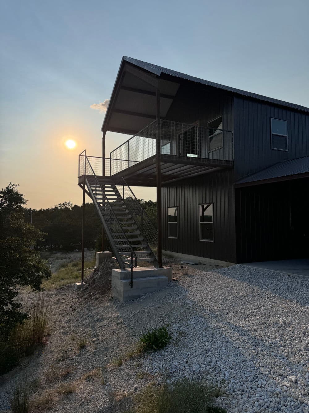 Modern black home with metal stairway and sunset backdrop, featuring a gravel driveway.