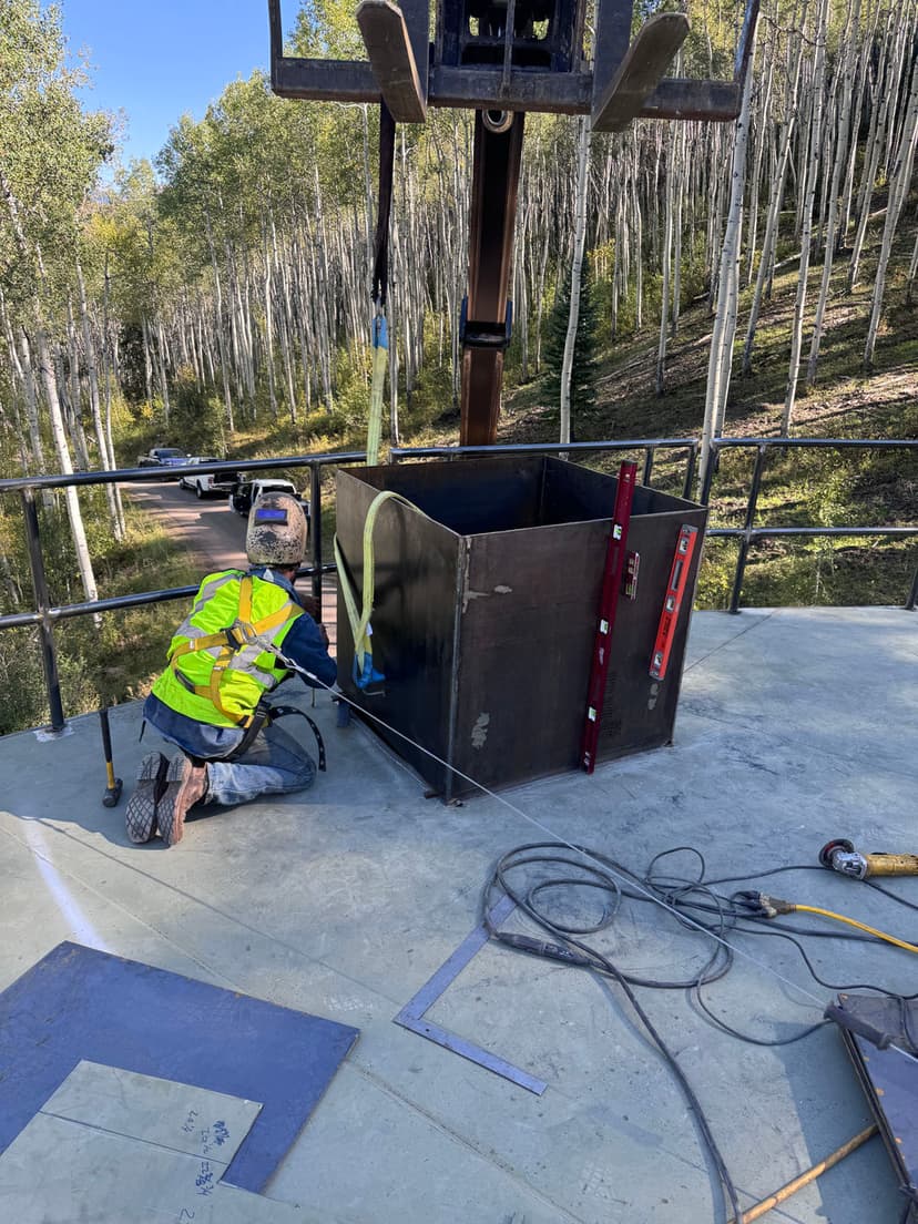 Worker securing a metal box on a construction site surrounded by aspen trees.