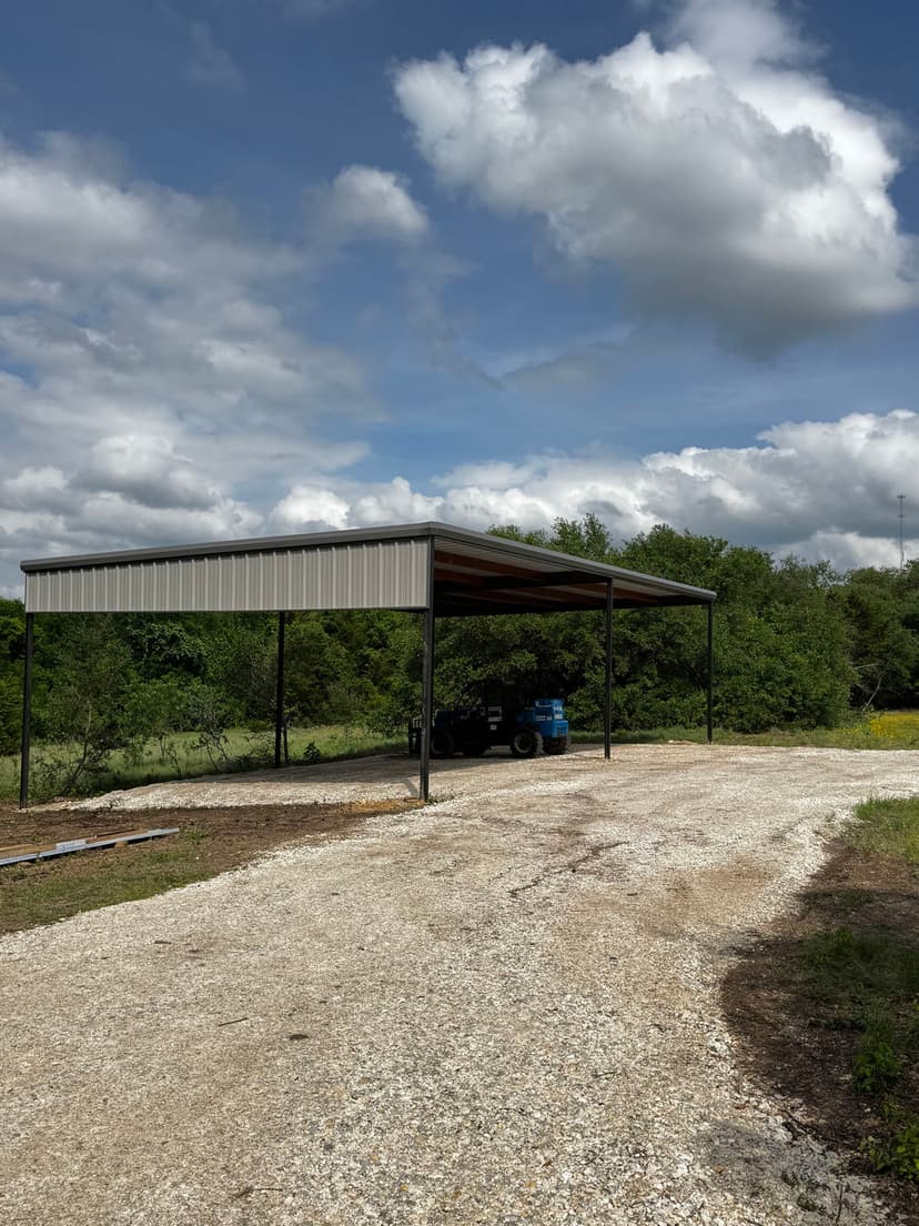 Metal carport structure on a gravel driveway surrounded by greenery and cloudy sky.