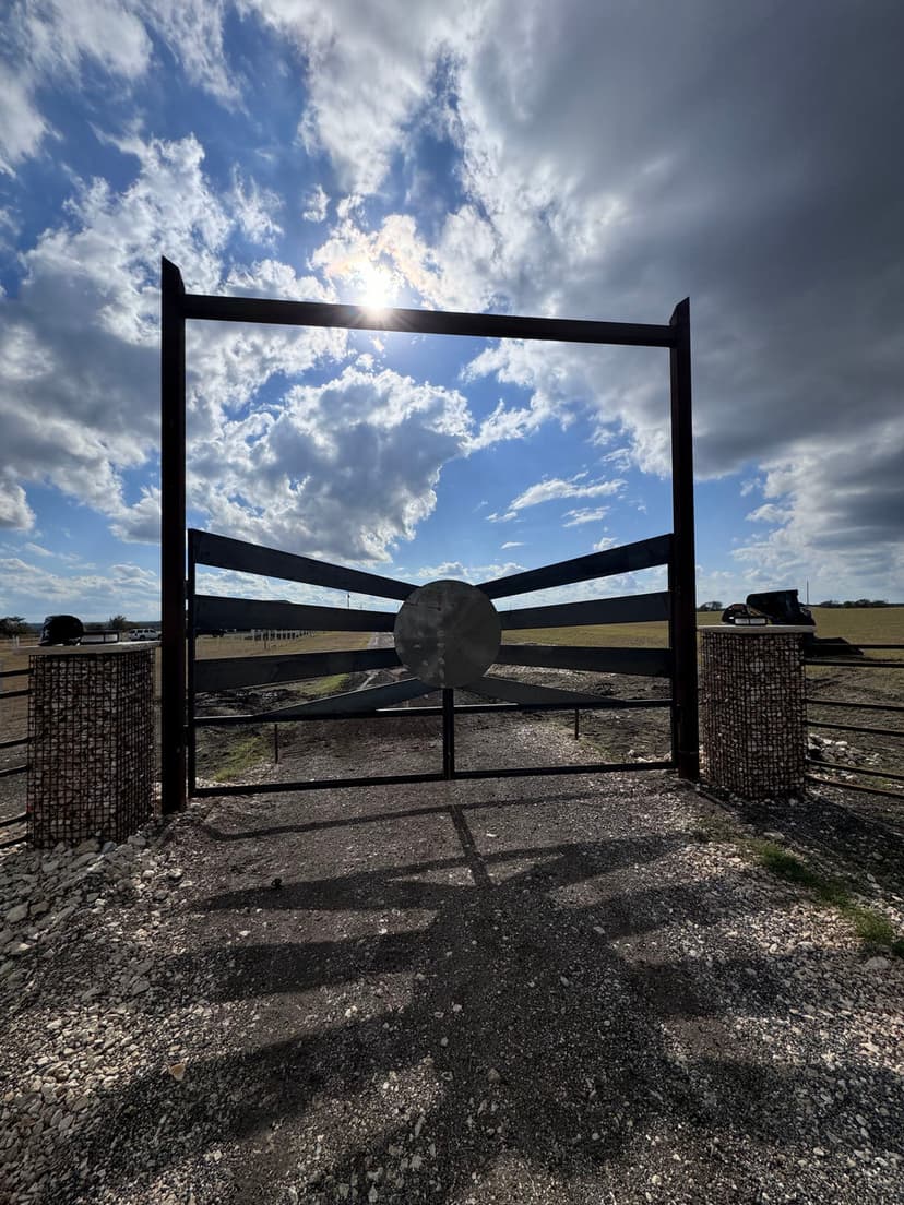 Large metal gate with a circular centerpiece against a cloudy sky in rural landscape.